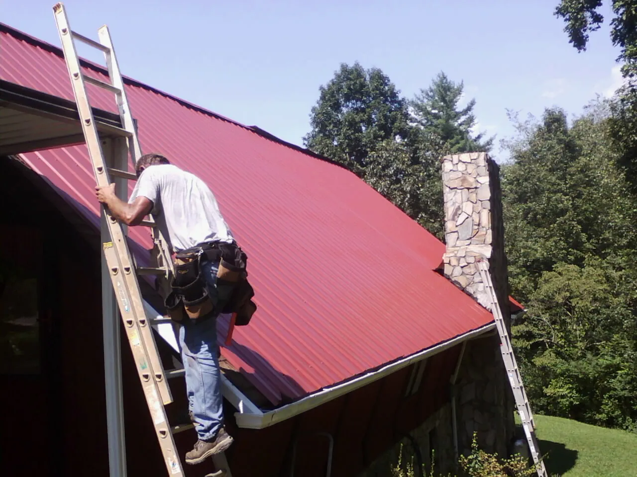 Roofer on a ladder beside a red metal roof and stone chimney — Soffit & Fascia Repair by Roofing Brothers in Zieglerville